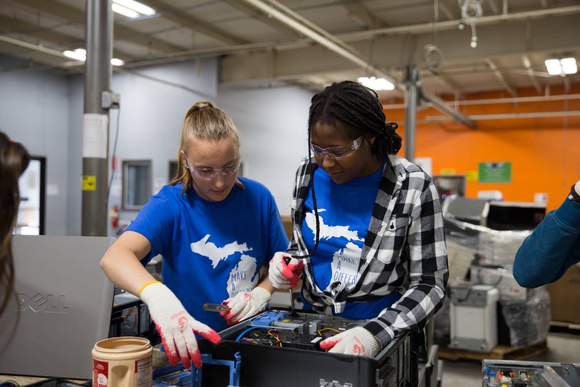 Two students wearing blue shirts working to take a part of a computer wearing safety clear goggles and white gloves